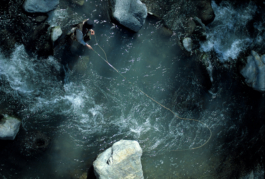 image from above of a person fly fishing with the rod and wire in hand, along a stretch of river with a fast flow, also some rocks. Ross Woodhall - Action & Lifestyle Photography