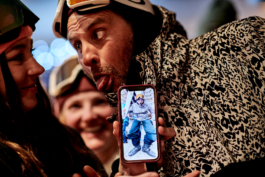 three skiers, a woman showing a picture of the man on her phone in his ski gear. He is looking at her, sticking his tongue out. Ross Woodhall - Action & Lifestyle Photography