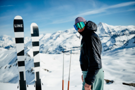 Man looking towards the camera, wearing winter clothes with googles on. Beside him is a pair of skis and poles pushed upright into the snow. Ross Woodhall - Action & Lifestyle Photography