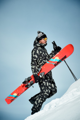 A man in a one piece snowsuit walking up the slope with a monoski and poles. Ross Woodhall - Action & Lifestyle Photography