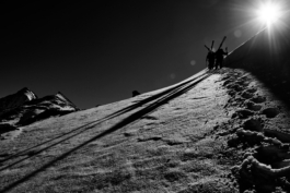 Black and white image. two skiers hiking up the mountain with skis over there shouldes to the right of the image, casting a shadow to the left with the sun. Leaving foot prints in the snow. Ross Woodhall - Action & Lifestyle Photography