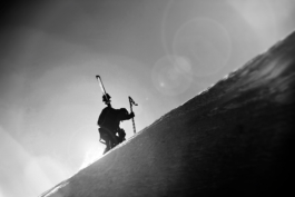 Black and white image of a skier hiking with skis over left shoudler, shot from below. Ross Woodhall - Action & Lifestyle Photography