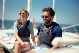Couple sat on a small sailing boat wearing a life vets with sunglasses on along the coastline with other boats in the background. Ross Woodhall - Action & Lifestyle Photography