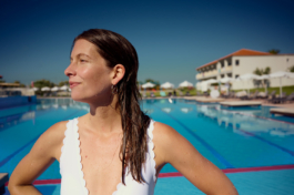 A woman just out of the pool wearing a white swimming costume, a large outdoor pool with sun loungers and umbrellas in the background. Ross Woodhall - Action & Lifestyle Photography