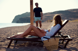 Woman relaxing, reading a book on a sun lounge on the beach with a man in the background strolling up towards her with a smile. Ross Woodhall - Action & Lifestyle Photography