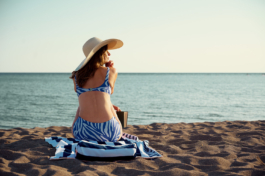 A woman sat on a sandy beach on a stripped towel taking a pause from reading her book, wearing an oversized hat, looking towards the sea. Ross Woodhall - Action & Lifestyle Photography