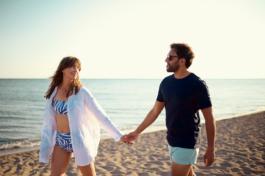 A man and woman holding hands walking along the coastline with a calm sea on a sandy beach. Gazing at each other. Ross Woodhall - Action & Lifestyle Photography