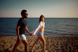 A man and woman holding hands walking along the coastline with a calm sea on a sandy beach. Ross Woodhall - Action & Lifestyle Photography