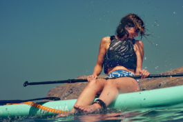 Woman sat on a paddle board, with a life jacket on, with the paddle across her, shielding herself from being splashed. Ross Woodhall - Action & Lifestyle Photography