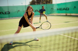 A couple playing doubles outside at tennis with the woman wearing black tennis outfit and laughing towards the camera. Ross Woodhall - Action & Lifestyle Photography