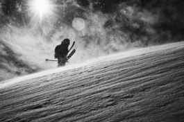 Black and white image of a skier holding each ski in both hands, walking up hill, with the sun and snow particles in the sky. Ross Woodhall - Action & Lifestyle Photography