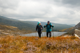 a hiking couple walking through the rough grass and heather with a lake in the background. Ross Woodhall - Action & Lifestyle Photography