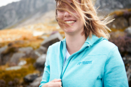 Woman hiking, closing her zip on her hoody, smiling with her hair caught in the wind across her face. Ross Woodhall - Action & Lifestyle Photography