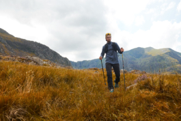 Man hiking with poles through the long grass with hills in the background. Ross Woodhall - Action & Lifestyle Photography
