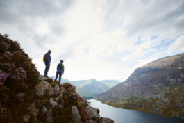 couple paused while hiking, looking out on a rocky path with heather towards the lake with hills in the background. Ross Woodhall - Action & Lifestyle Photography