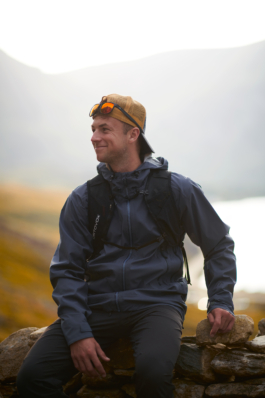 man sat on a stone wall, wearing a jacket, backpack. with a cap and sunglasses on, with lake and mountain in the background. Ross Woodhall - Action & Lifestyle Photography