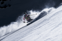 female skier wearing a pink jacket, turning off piste with poles out with flicked up snow getting caught by the wind. Ross Woodhall - Action & Lifestyle Photography