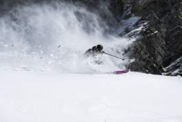 skier off piste mid turn with pole out stretched with snow flicked up which the wind has caught. Background is base of a rocky area. Ross Woodhall - Action & Lifestyle Photography