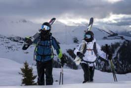 two skiers with skis over their shoulders walking towards the camera with piste, lifts and trees in background Ross Woodhall - Action & Lifestyle Photography