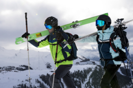 close up view of two skiers walking with skis over their shoulders, one female, one male with piste runs and lifts in background with trees on the mountain. Ross Woodhall - Action & Lifestyle Photography