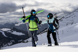 two skiers one female, one male walking with skis over their shoulders in fresh snow with piste in the background, chatting to each other. Ross Woodhall - Action & Lifestyle Photography
