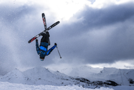 Skier mid air upside down with skis crossed into an X with mountains and lift system with clouds in the sky, looking moody. Ross Woodhall - Action & Lifestyle Photography