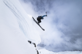 Skier off piste just launched off a small cornice with mountain and lifts in the background on a cloudy day Ross Woodhall - Action & Lifestyle Photography