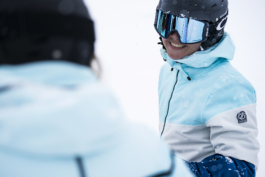 view from over the shoulder of a skier towards another skier who is smiling back, wearing helmet, googles and a jacket in the snow. Ross Woodhall - Action & Lifestyle Photography